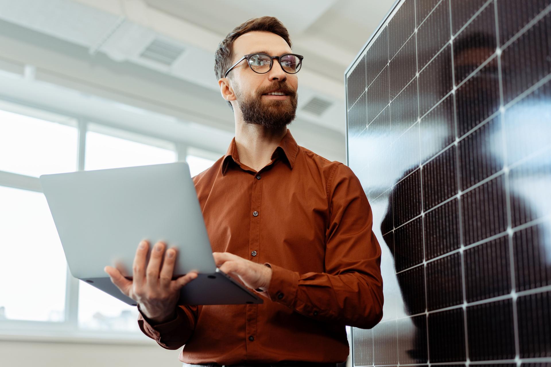 Handsome smiling man, developer holding laptop planning startup working with solar panel in office